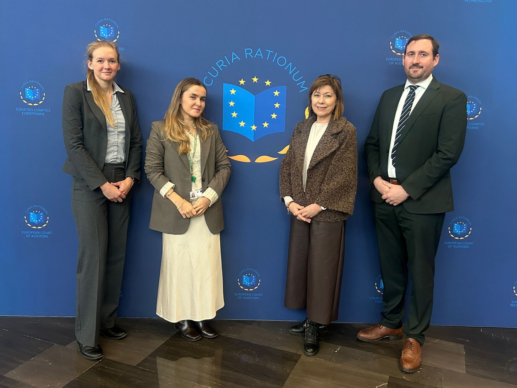 Während ihres Aufenthalts erhielt Andrea wertvolle Einblicke in die Aufgabenbereiche, Abläufe und die Arbeitsweise des Kabinetts von Helga Berger, Mitglied des Europäischen Rechnungshofes. Auf dem Foto stehen von links nach rechts: Andrea Preinfalk, Sina Prokop (Trainee), Silvia Janik (Head of Cabinet) und Franz Ebermann (Attaché) vor einem blauen Hintergrund mit einem Logo mit gelben Sternen und einem blauen Buch sowie der Überschrift Curia Rationum.
