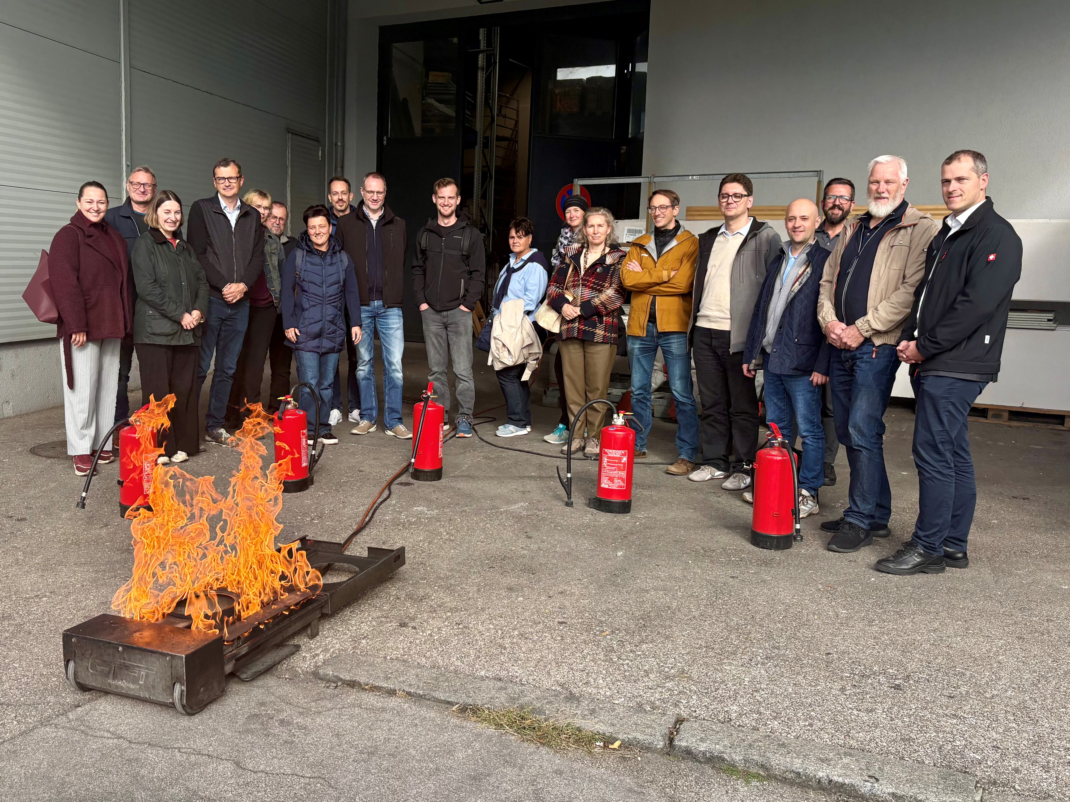 Gruppenfoto mit mehreren Menschen im Zuge der Besichtigung der Brandverhütungsstelle OÖ in Linz vor der Lagerhalle und einer Feuerquelle